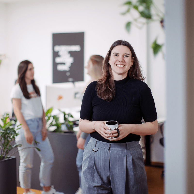 Eine Person steht l&auml;chelnd mit einer Kaffeetasse im Vordergrund, w&auml;hrend im Hintergrund drei Kolleginnen miteinander sprechen.
