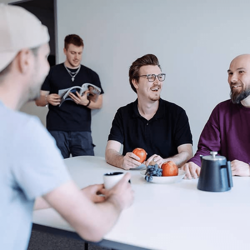 Vier Teammitglieder von mindtwo im Gespr&auml;ch am Tisch, mit Kaffee und Obst, im Hintergrund liest jemand eine Zeitschrift